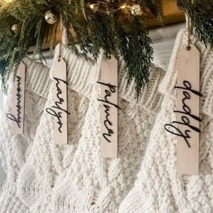 Puede incluir: Cuatro calcetines de punto blanco con etiquetas de madera que cuelgan de una guirnalda de vegetación con luces y campanas. Las etiquetas están personalizadas con los nombres "Mommy", "Harlyn", "Payton" y "Daddy".