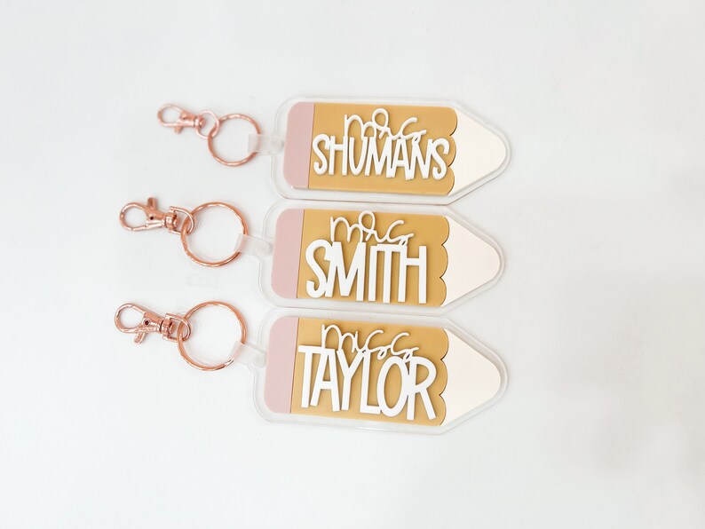 May include: Three gold pencil-shaped keychains with pink accents and white text. The keychains read "Mrs. Shumans", "Mrs. Smith", and "Mrs. Taylor".