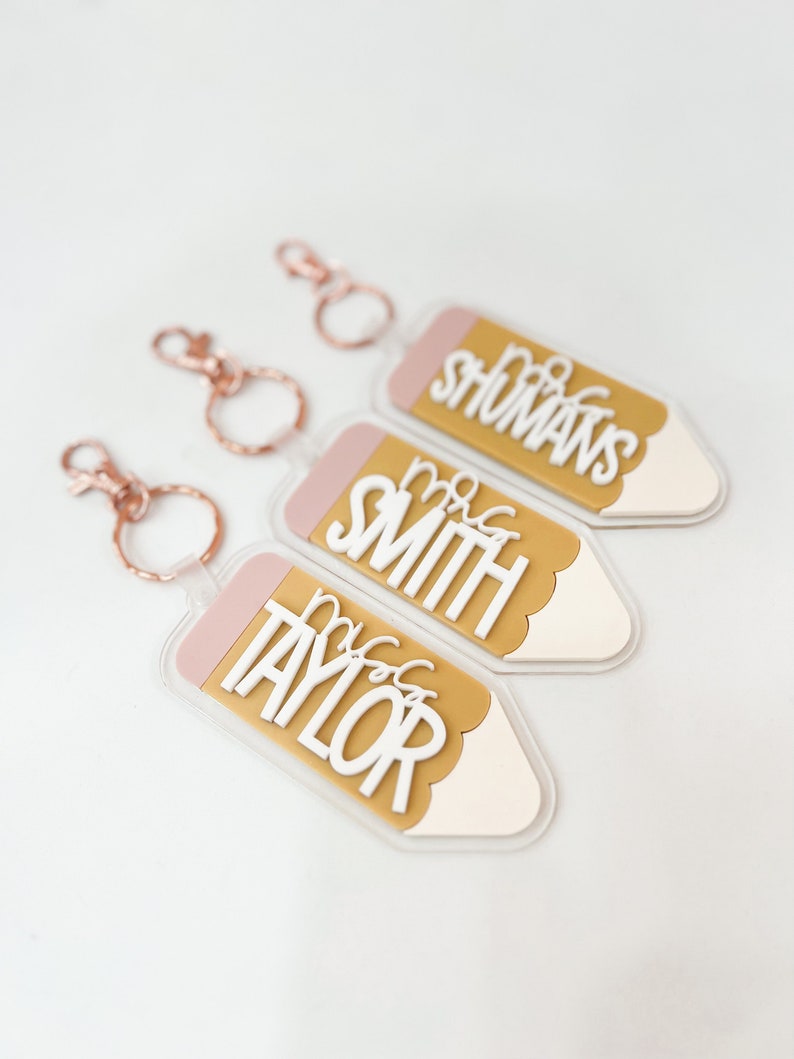 May include: Three gold pencil-shaped keychains with pink accents and white lettering. The keychains read "Mrs. Taylor", "Mrs. Smith", and "Mrs. Shomans".