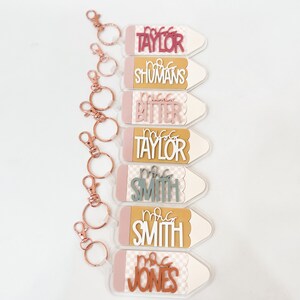 May include: Seven pink and white pencil-shaped keychains with gold key rings. Each keychain has a different teacher's name on it, including "Mrs. Taylor", "Mrs. Shumans", "Mrs. Bitter", "Mrs. Taylor", "Mrs. Smith", "Mrs. Smith", and "Mrs. Jones".