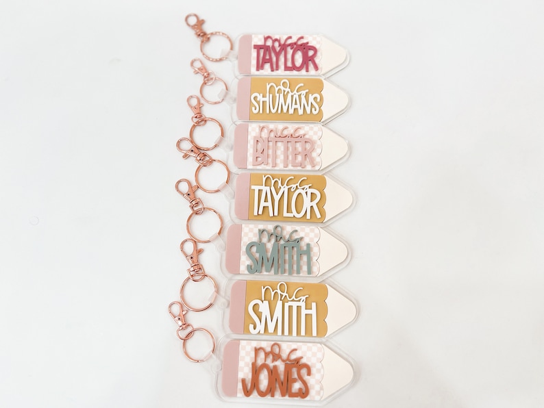 May include: Seven pink and white pencil-shaped keychains with gold key rings. Each keychain has a different teacher's name on it, including "Mrs. Taylor", "Mrs. Shumans", "Mrs. Bitter", "Mrs. Taylor", "Mrs. Smith", "Mrs. Smith", and "Mrs. Jones".