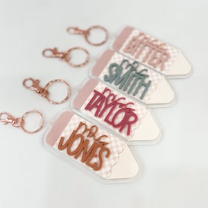 May include: Four clear acrylic pencil-shaped keychains with different teacher names. The keychains have a pink and white checkered pattern and a scalloped edge. The names are "Mrs. Bitter", "Mrs. Smith", "Mrs. Taylor", and "Mrs. Jones".