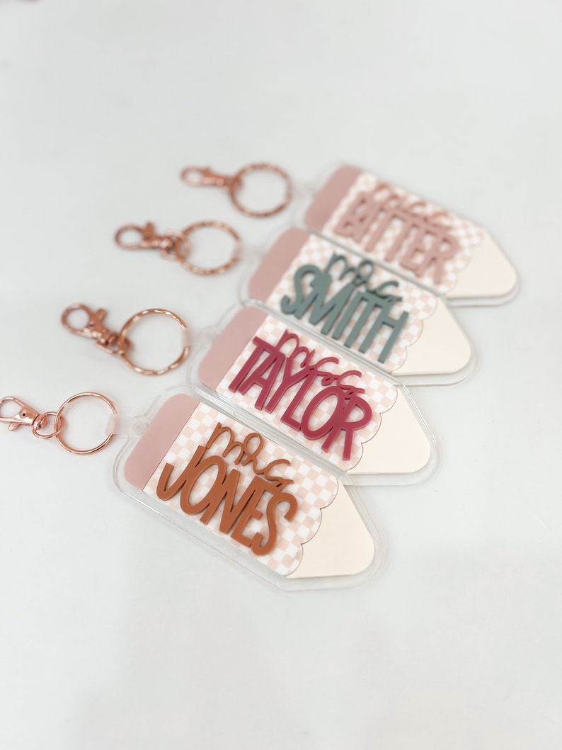 May include: Four clear acrylic pencil-shaped keychains with different teacher names. The keychains have a pink and white checkered pattern and a scalloped edge. The names are "Mrs. Bitter", "Mrs. Smith", "Mrs. Taylor", and "Mrs. Jones".