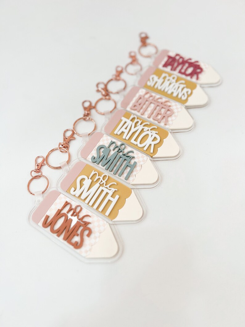 May include: A set of six pencil-shaped keychains with different teacher names. Each keychain is made of clear acrylic with a pink and yellow patterned background. The names are written in white and gold letters.