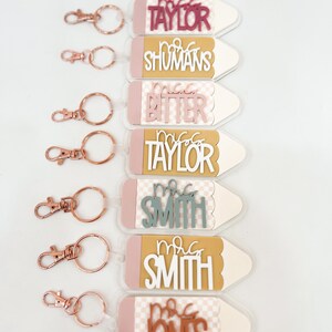 May include: Set of six pencil-shaped keychains with different teacher names. Each keychain has a pink and white checkered pattern, a gold top, and a rose gold key ring.