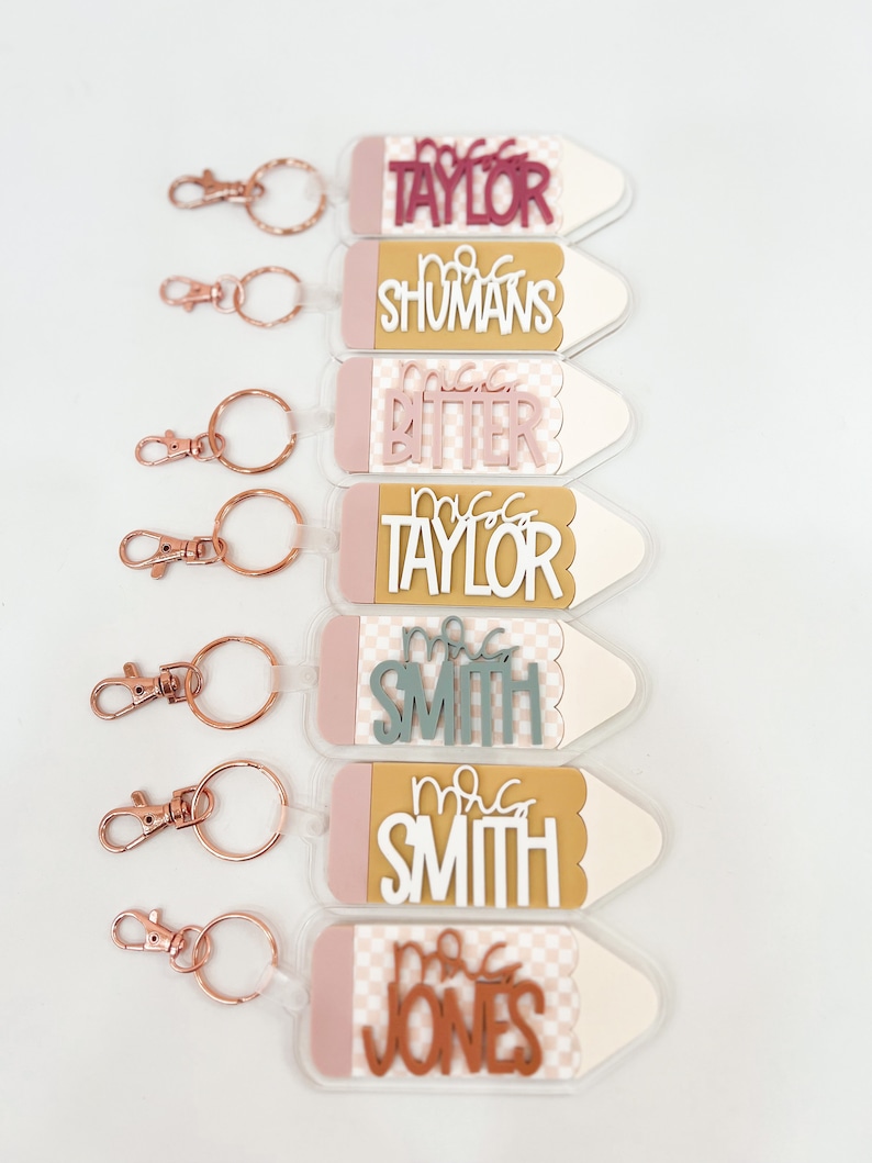 May include: Set of six pencil-shaped keychains with different teacher names. Each keychain has a pink and white checkered pattern, a gold top, and a rose gold key ring.