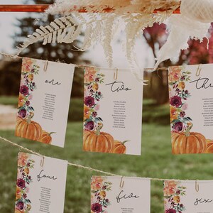 May include: Six white cards with floral and pumpkin designs are clipped to a string. Each card is numbered one through six and includes a list of names. The cards are decorated with watercolor flowers in shades of orange, burgundy, and yellow.