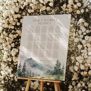 May include: A wedding seating chart on a wooden easel, featuring the text "OUR FAVORITE PEOPLE" and a watercolor mountain scene. The chart is surrounded by white roses, creating an elegant and natural aesthetic. The names "Olivia & Jason" are also visible.