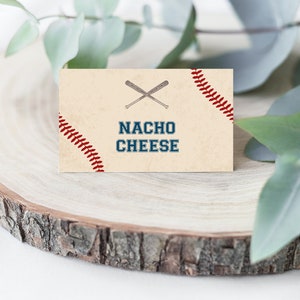 May include: A baseball-themed card with the words "NACHO CHEESE" in blue, vintage-style lettering. The card features crossed baseball bats and red stitching, resembling a baseball. The card is set on a wooden slice.