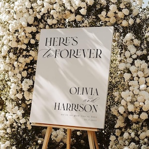 May include: A white sign on a wooden easel with the text "HERE'S TO FOREVER" and the names "OLIVIA and HARRISON". The sign is surrounded by white flowers and greenery, perfect for a wedding or event.