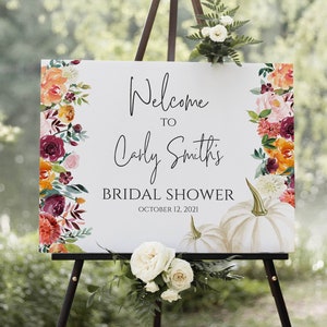 Puede incluir: Un letrero blanco sobre un caballete negro con el texto "Welcome to Carly Smith's Bridal Shower October 12, 2021". El letrero está decorado con arreglos florales en tonos naranja, burdeos y blanco, con dos calabazas blancas.