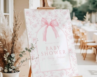 May include: A baby shower welcome sign on a wooden easel. The sign features a pink floral pattern and a pink bow. Text reads "Welcome to the Baby Shower for Ashley Anderson" and the date.