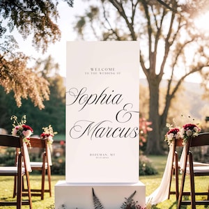 Pu&ograve; includere: Un cartello di benvenuto per matrimoni bianco con la scritta "WELCOME TO THE WEDDING OF Sophia & Marcus" in elegante corsivo. Il cartello include anche il luogo e la data. Il cartello &egrave; esposto all'aperto.
