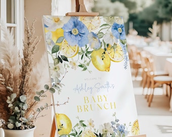May include: A welcome sign on a wooden easel, featuring a floral design with lemons and blue flowers. The sign reads "Welcome to Ashley Smith's Baby Brunch" in elegant script and bold text. The background is blurred, suggesting an outdoor event.