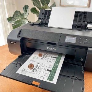 May include: A black Canon PRO-300 printer with a white sheet of paper in the tray. The printer is printing a green and white football-themed invitation with the text "WESTON'S Tailgate Dinner" and "SEPT. 17 8:30PM".