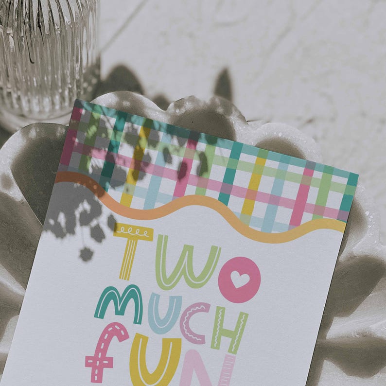 May include: A greeting card with the words "TWO MUCH FUN" in colorful, playful lettering. The card features a pastel plaid pattern at the top and is resting on a white, textured surface.