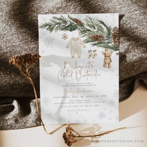 May include: Baby shower invitation with the text "Baby it's Cold Outside". The invitation features a watercolor illustration of a baby onesie, socks, and a teddy bear hanging from pine branches. The invitation includes the name Carly Anderson and event details.