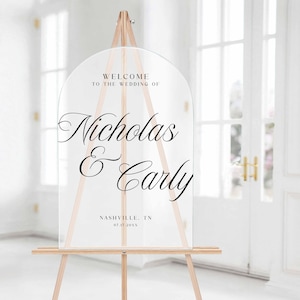 Pu&ograve; includere: Cartello di benvenuto per matrimoni in acrilico trasparente su un cavalletto di legno. Il cartello ha una parte superiore ad arco e presenta un'elegante scritta nera che recita "Benvenuti al matrimonio di Nicholas & Carly" e il luogo e la data.