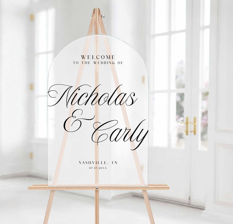 Pu&ograve; includere: Cartello di benvenuto per matrimoni in acrilico trasparente su un cavalletto di legno. Il cartello ha una parte superiore ad arco e presenta un'elegante scritta nera che recita "Benvenuti al matrimonio di Nicholas & Carly" e il luogo e la data.