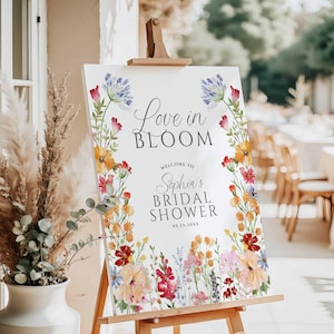 May include: A white sign on a wooden easel with the text "Love in Bloom" and "Sophia's Bridal Shower." The sign is decorated with colorful watercolor flowers in shades of pink, yellow, orange, and blue. The date 05.25.20xx is also included.