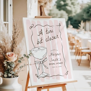 May include: A pink and white sign with the text "A tini bit closer" and a drawing of a cocktail glass. The sign also says "Welcome to Harper Smith's Bridal Shower May 25, 20xx".