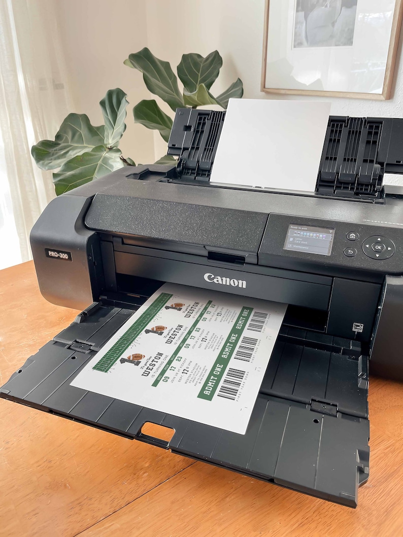 May include: A black Canon printer with a white paper tray. The printer is printing out a sheet of paper with green and white tickets that say "Weston" and "Admit One".