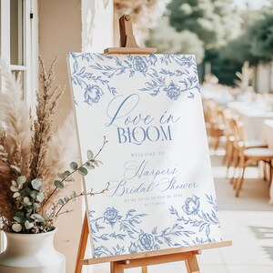May include: A white sign on a wooden easel with blue floral accents and text. The sign reads "Love in Bloom" and welcomes guests to "Harper's Bridal Shower" with the date 07.17.2011. Decorative floral border.