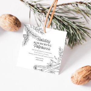 Peut inclure: Une étiquette cadeau imprimable blanche avec du texte noir et des illustrations. Le texte indique "Holiday Simmering Potpourri" et comprend des instructions pour faire du pot-pourri. L'étiquette est attachée à un cordon en cuir brun et est suspendue à une branche de pin.