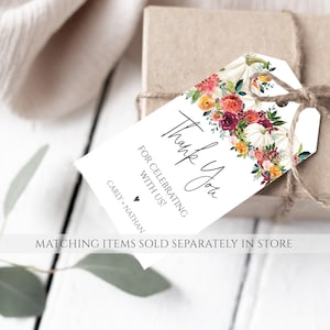 Op de afbeelding: Een wit cadeaulabel met bloemen- en pompoenaccenten, vastgebonden aan een bruine doos met touw. Op het label staat "Thank You" en "For Celebrating With Us!" met de namen "Carly + Nathan" eronder. Het label is versierd met aquarelbloemen.