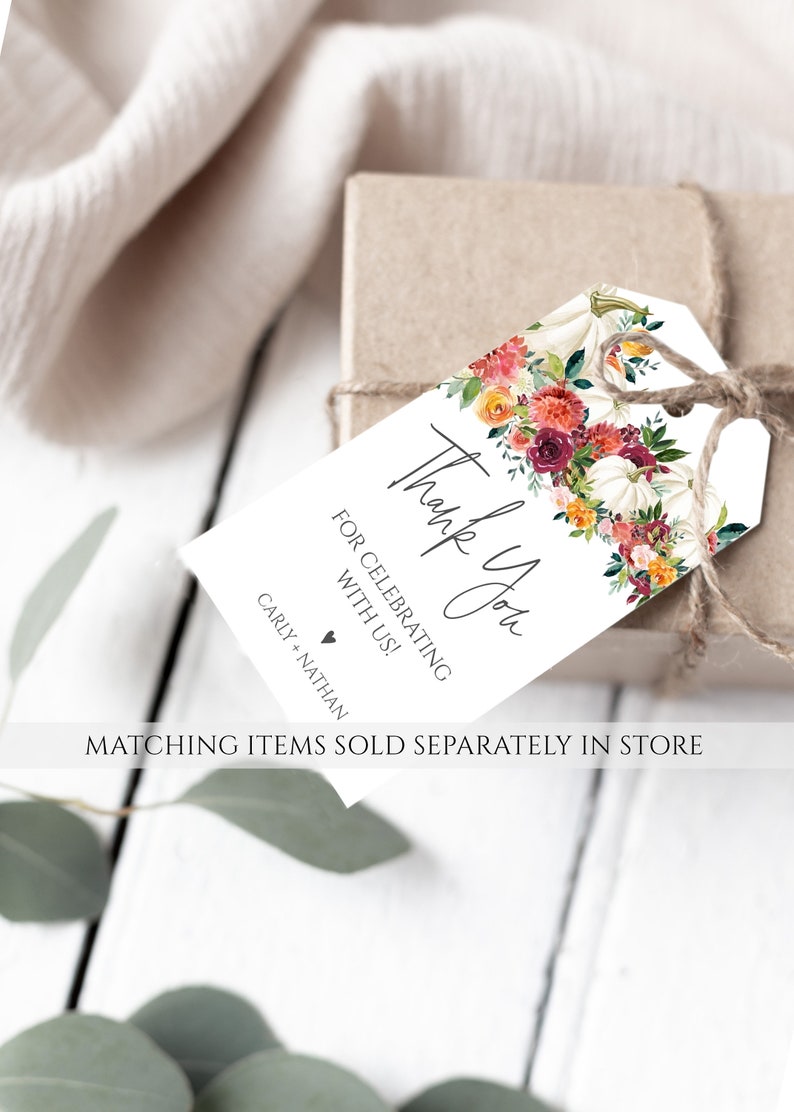 Op de afbeelding: Een wit cadeaulabel met bloemen- en pompoenaccenten, vastgebonden aan een bruine doos met touw. Op het label staat "Thank You" en "For Celebrating With Us!" met de namen "Carly + Nathan" eronder. Het label is versierd met aquarelbloemen.