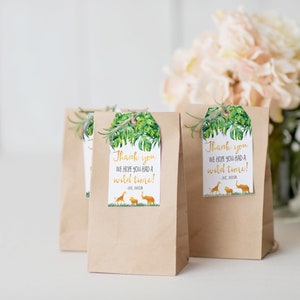 Pode incluir: Sacos de presente de papel kraft com etiquetas com tema de selva. As etiquetas apresentam um design de folha tropical, o texto "Thank you, we hope you had a wild time!" e silhuetas de uma girafa, le&atilde;o e elefante. Um ramo de vegeta&ccedil;&atilde;o est&aacute; anexado.