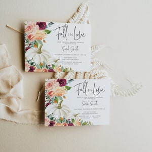 Op de afbeelding: Twee witte kaarten met de tekst "Fall in Love" worden getoond. De kaarten bevatten bloemstukken met witte pompoenen, rozen en groen. Een cr&egrave;mekleurig lint en varen zijn ook aanwezig, waardoor een herfstthema bruidsdouche-uitnodiging ontstaat.
