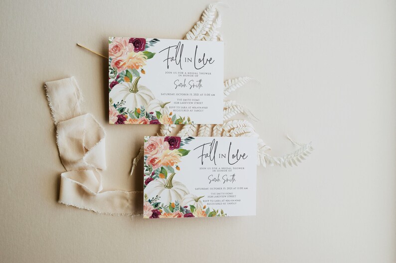 Op de afbeelding: Twee witte kaarten met de tekst "Fall in Love" worden getoond. De kaarten bevatten bloemstukken met witte pompoenen, rozen en groen. Een cr&egrave;mekleurig lint en varen zijn ook aanwezig, waardoor een herfstthema bruidsdouche-uitnodiging ontstaat.