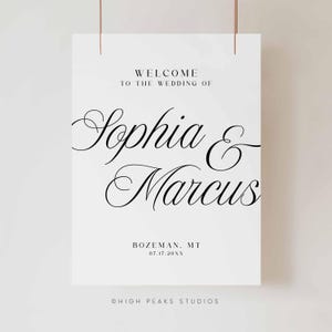 Pu&ograve; includere: Un cartello di benvenuto per matrimoni bianco con scritte nere. Il cartello recita "WELCOME TO THE WEDDING OF Sophia & Marcus" con la localit&agrave; e la data sottostanti. Il cartello &egrave; sospeso da due sottili fili color rame.