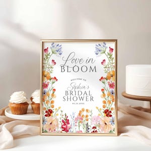 May include: A framed sign with the words "Love in Bloom" and "Welcome to Sophia's Bridal Shower" in elegant script. The sign is decorated with colorful floral arrangements in shades of pink, orange, and blue. Cupcakes and a cake are in the background.
