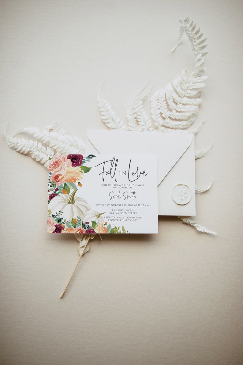 Op de afbeelding: Uitnodiging voor een bruidsdouche met de tekst "Fall in Love" en details voor de douche van Sarah Smith. De uitnodiging heeft een bloemen- en pompoenontwerp. Een envelop en decoratieve witte varen zijn ook aanwezig.