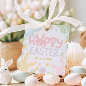 Puede incluir: Una etiqueta de regalo de Pascua rosa y blanca con un conejito y el texto "Hoppy Easter" y "With love, Harper". La etiqueta está decorada con flores y conejos pastel.