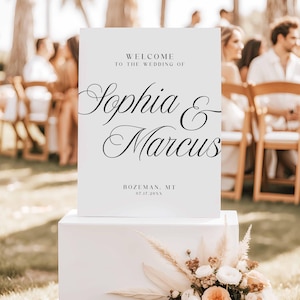 Pu&ograve; includere: Un cartello di benvenuto per matrimoni bianco con la scritta nera che recita "Benvenuti al matrimonio di Sophia & Marcus". Il cartello include anche il luogo e la data. Il cartello &egrave; esposto su un piedistallo bianco con composizioni floreali.