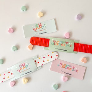May include: Four Valentine's Day themed slap bracelets with colorful designs and text. The bracelets are on a white surface with candy hearts scattered around. The text on the bracelets reads "Oh Snap! Happy Valentine's Day!" and "Slappy Valentine's Day!"