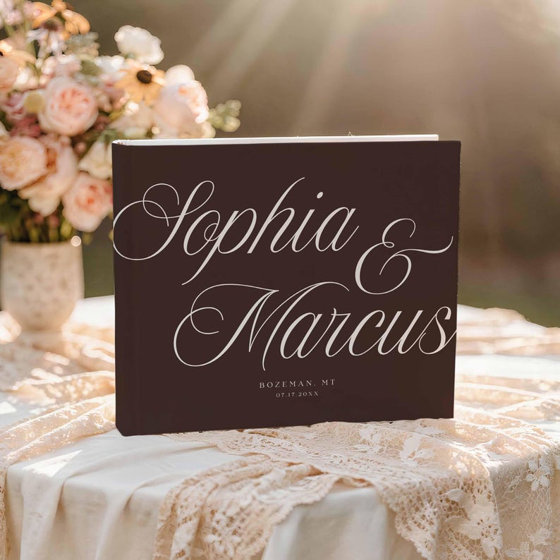 May include: A dark brown rectangular photo album with white script text reading Sophia & Marcus, followed by the location Bozeman, MT and the date 07.17.20XX. The cover features a clean, matte finish with elegant, flowing typography centered on the front.