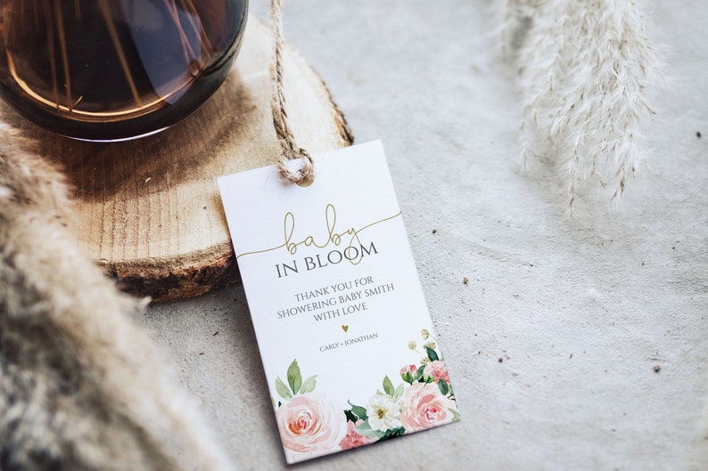 May include: A white baby shower favor tag with gold lettering that reads "baby in bloom". The tag has a floral design with pink and white flowers and green leaves. The text below the title reads "Thank you for showering baby Smith with love" and is followed by the names "Carly + Jonathan".