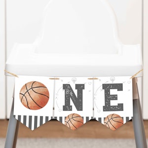 May include: A banner with the word "ONE" for a first birthday celebration. The banner features basketball graphics and a black and white striped pattern. The banner is hanging on a white high chair.