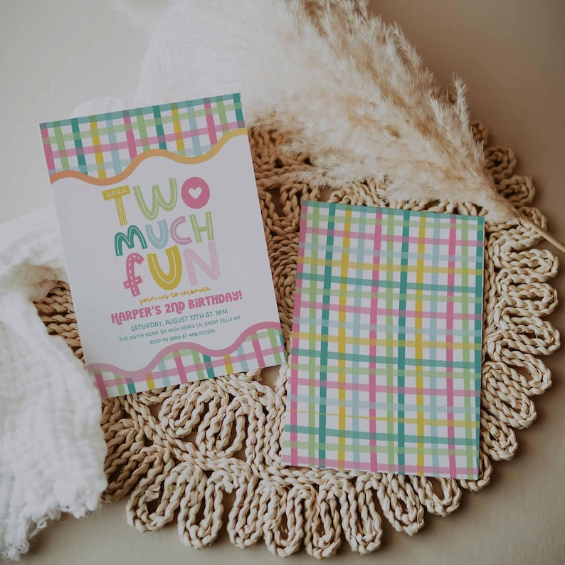 May include: Two birthday invitations with a colorful plaid pattern and the words "TWO MUCH FUN." One invitation has the text "Harper's 2nd Birthday!" The invitations are on a woven placemat with a white cloth and dried plants.