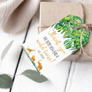 Pode incluir: Uma etiqueta de presente com tema de selva, com folhas tropicais verdes e texto dourado que diz "Obrigado, esperamos que voc&ecirc; tenha se divertido!" com silhuetas de animais dourados. A etiqueta est&aacute; amarrada a uma caixa marrom com barbante.