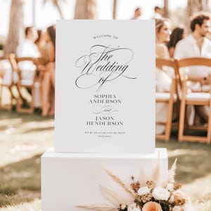 May include: A white rectangular wedding welcome sign with black text that reads Welcome to The Wedding of Sophia Anderson and Jason Henderson, We're so glad you're here, and 05.25.20XX. The sign is displayed on a white pedestal with floral decor.