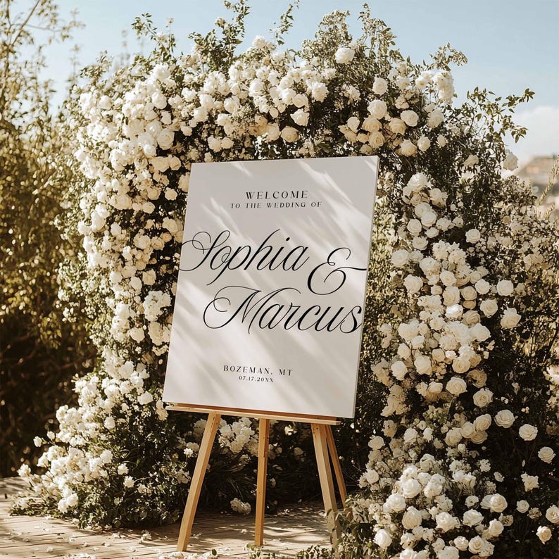 May include: A white wedding welcome sign on a wooden easel, with the names Sophia & Marcus in elegant script. The sign is surrounded by a lush backdrop of white roses and greenery, with the text "Welcome to the Wedding of" above the names and the location and date below.