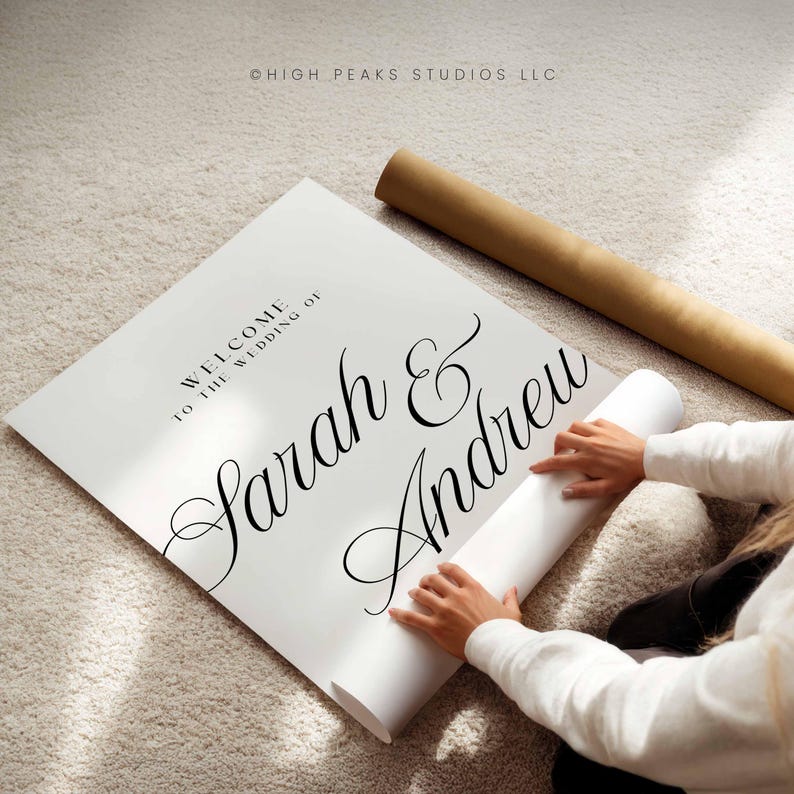 Pu&ograve; includere: Un cartello nuziale bianco con i nomi "Sarah & Andrew" in elegante scrittura. Il cartello recita "Benvenuti al matrimonio di" in alto. Sono visibili anche un poster arrotolato e un tubo di cartone marrone.