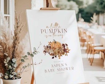 May include: A white sign on a wooden easel with the text "a little PUMPKIN is on the way!" in brown script. A pumpkin and floral arrangement is below the text. The sign welcomes guests to Aspen's Baby Shower.