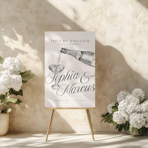 May include: A rectangular white sign on a gold easel featuring black text that reads THEY'RE ENGAGED LET'S CELEBRATE, Sophia & Marcus, and ROZEMAN, MI 07.17.20XX. The design includes a black and white sketch of a champagne bottle pouring into a glass.
