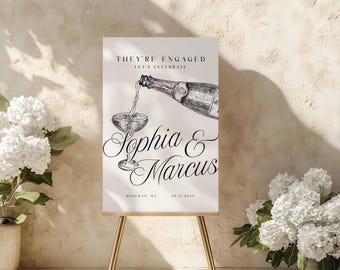 May include: A rectangular white sign on a gold easel featuring black text that reads THEY'RE ENGAGED LET'S CELEBRATE, Sophia & Marcus, and ROZEMAN, MI 07.17.20XX. The design includes a black and white sketch of a champagne bottle pouring into a glass.